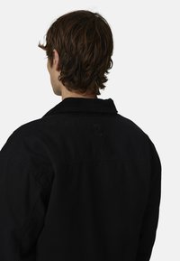 Giacca di velluto a costine nera con colletto, cuciture visibili e un logo circolare sulla parte superiore della schiena. Il tessuto ha un aspetto texture.