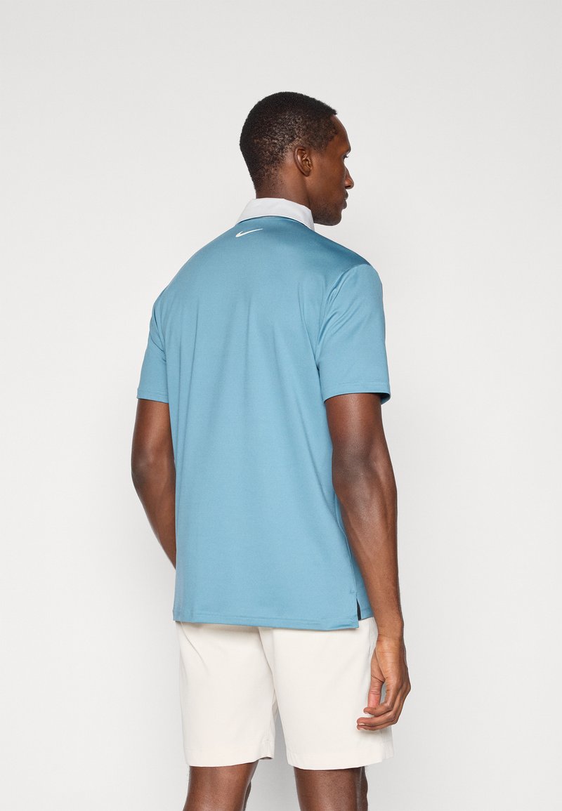 Lys blå polo skjorte med et sort logo på ryggen, korte ærmer og en kontrasterende hvid krave. Båret med beige shorts.