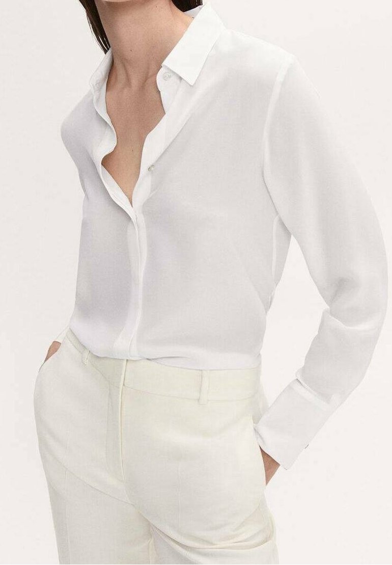 Femme portant une chemise blanche boutonnée avec les premiers boutons défaits et un pantalon taille haute couleur crème, les mains dans les poches.