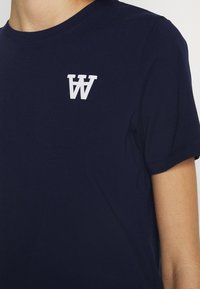 Tmavě modré tričko s kulatým výstřihem vyrobené z bavlny. Má malé bílé logo na hrudi a krátké rukávy. Hladká textilie.