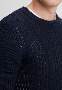 Marineblauer gestrickter Pullover aus strukturiertem Garn mit Kabelmuster und geripptem Rundhalsausschnitt, der komplexe Details und offene Bereiche aufweist.
