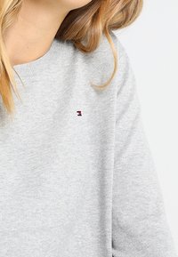 Světle šedá mikina z měkké látky s kulatým výstřihem. Na hrudi je malé logo v červené, bílé a modré barvě.