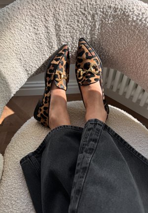 Luipaardprint loafers met een spitse neus en gouden hardwaredetails, rustend op een gestructureerde lichtgekleurde stoel met zichtbaar zwart denimbroek.