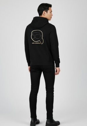 Mann trägt schwarzen Hoodie und Hose, steht seitlich, auf der Rückseite des Hoodies ist das Karl Lagerfeld-Logo mit stilisiertem Gesicht und Brille zu sehen.