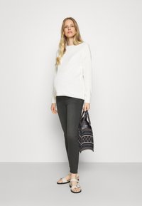 Maglione bianco lavorato a maglia con maniche lunghe, jeans skinny grigi e sandali beige. Tiene una borsa a motivo blu scuro. Tessuto liscio e design semplice.