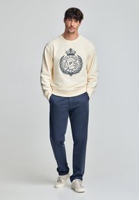 Felpa crema con logo e emblema blu, abbinata a pantaloni blu. L'outfit presenta una vestibilità comoda e un tessuto testurizzato. Sneakers bianche.