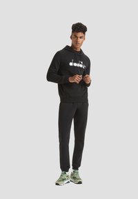 Felpa nera con logo bianco, pantaloni della tuta neri abbinati e sneakers con accenti verdi. Tessuto morbido e vestibilità rilassata, progettata per il comfort.