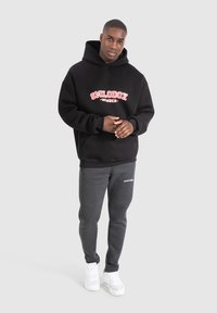 Černá mikina s červeným nápisem "SMILODOX MEMBER", v kombinaci se šedými tepláky s bílým logem a bílými teniskami. Model stojí, ruce uvolněné.