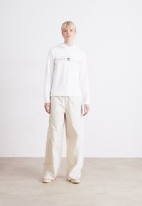 Hvit hettegenser med grå tekst og logo, kombinert med lyse beige vide bukser. Modellen står foran en lys, nøytral bakgrunn.