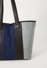 Sac fourre-tout avec des poignées en cuir noir, un panneau en denim bleu foncé et un côté en denim clair, avec une forme rectangulaire structurée.