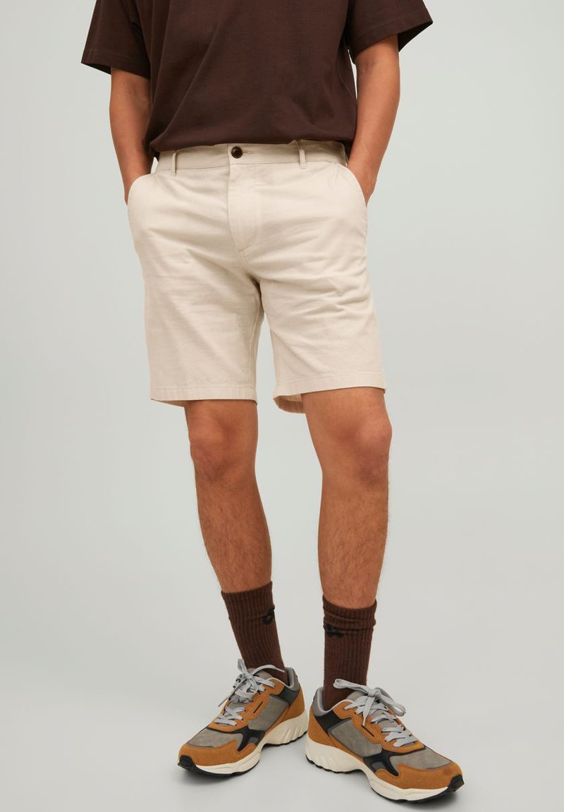 Shorts en coton beige avec poches latérales, fermeture à bouton et coupe ajustée. Associés à des baskets grises et brunes et des chaussettes marron.