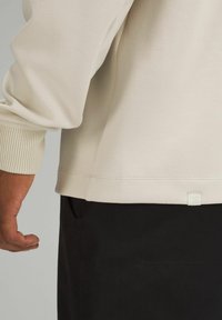 Beige Sweatshirt mit gerippten Bündchen und Saum, ausgestattet mit einem kleinen Logo-Tag am unteren Ende, kombiniert mit schwarzen Hosen. Glatte Textur.