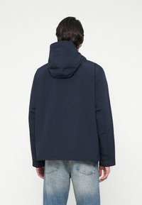 Egy férfi sötét tengeri kék kapucnis dzsekit és világoskék, kopott farmernadrágot visel, háttal állva egy sima fehér háttér előtt.