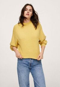 Pull jaune tricoté avec un col rond et une texture côtelée. Coupe ample avec des poignets retroussés, associé à un jean bleu taille haute.