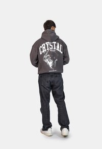 Felpa grigia scura con un grafico di un lupo bianco e il testo "CRYSTAL" sulla schiena. Indossa jeans neri strappati e sneakers bianche.