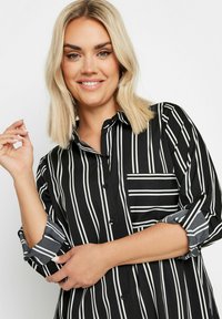 Camicia a righe verticali bianco e nere con bottoni davanti, maniche arrotolate e taschino sul petto. Tessuto dalla texture liscia.