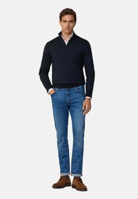 Marinblå stickad tröja med dragkedja i kragen, ljusblå skjorta under, blå jeans och bruna mockasko. Enkelt, figursytt design.