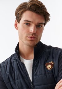 Marineblå pufjakke med lynlås foran og ribbet krave, med en tekstureret bjørnepatch på venstre bryst og en hvid t-shirt under.