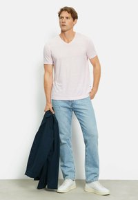 T-shirt rosa chiaro a V con maniche corte, abbinata a jeans azzurri e sneakers bianche, mentre tiene una giacca blu navy in una mano.