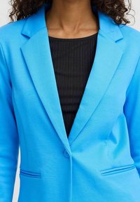 Kvinna i en ljusblå blazer över en svart ribbad topp, visad från halsen till midjan med lockigt hår delvis synligt.