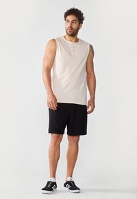 Helle beige ärmellose Bluse mit drei Knöpfen, kombiniert mit schwarzen Shorts. Die Sneakers bestehen aus schwarzem Stoff mit weißen Akzenten und Gummisohlen.