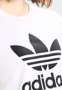 Camiseta de algodón blanca con un gran logo de trébol negro de Adidas y tres rayas. Mangas cortas y un cuello redondo. Textura suave.