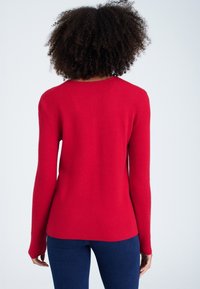 Röd stickad tröja med långa ärmar, rund halsringning och en åtsittande siluett. Slät yta och jämn färg. Bärs med mörkblå jeans.