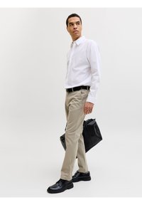 Άνδρας με λευκό πουκάμισο και μπεζ παντελόνι, κρατώντας μαύρη τσάντα, κοιτάζει στο πλάι, στέκεται μπροστά σε απλό φωτεινό φόντο.