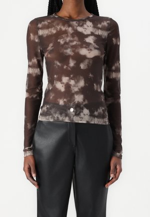 Femme portant un haut moulant à manches longues tie-dye marron et beige, associé à un pantalon en cuir noir taille haute, debout devant un fond blanc.