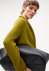 Maglione in maglia verde senape con superficie testurizzata e maniche ampie, abbinato a una grande borsa in pelle nera con dettagli di design angolari.