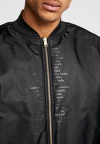 Svart bomberjacka med dragkedja, ribbad krage och präglad textdetaljering fram. Slät, glansig tygtextur.