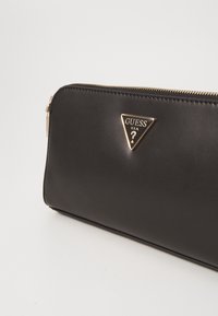 Schwarzes Lederportemonnaie mit rechteckiger Form. Verfügt über ein goldenes, dreieckiges Logo und einen Reißverschluss entlang der oberen Kante. Glatte Textur.