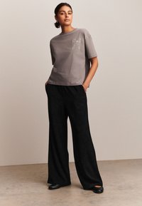 Camiseta gris de manga corta con un bordado de lazo en el pecho, combinada con pantalones negros de pierna ancha y zapatos negros estilo slip-on. Fondo liso.