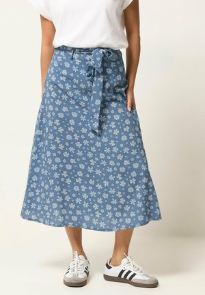 Mujer que lleva una falda azul hasta la rodilla con patrón floral blanco, cinturón atado, top blanco y zapatillas blancas con rayas negras.