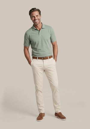 Lachende man staat met handen in de zakken, draagt een lichtgroen polo, beige broek, een bruine riem en bruine instapschoenen tegen een neutrale achtergrond.