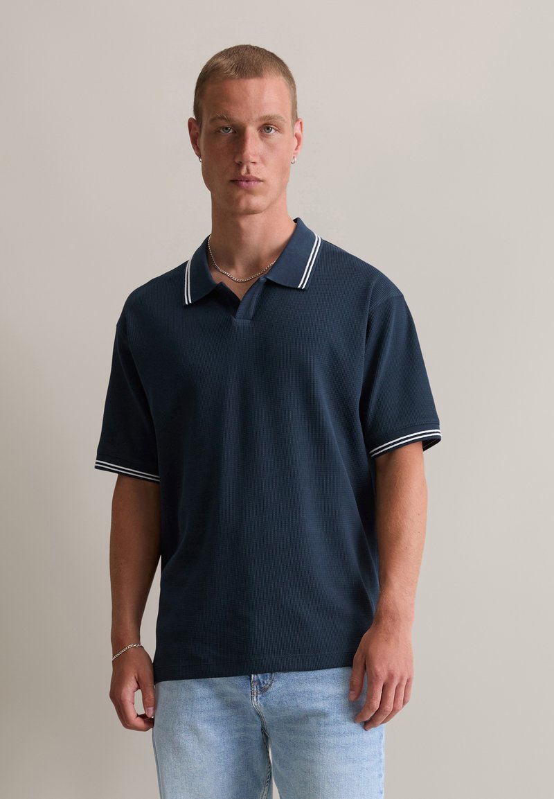 Granatowa koszulka polo z fakturowanej tkaniny, z kołnierzykiem w szpic oraz białymi paskami na kołnierzu i rękawach.