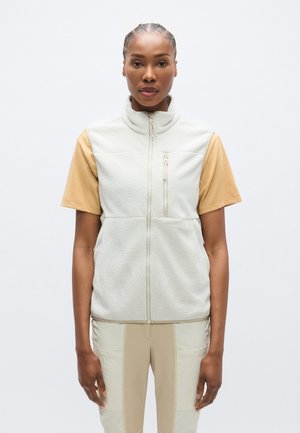 Fleecová vesta v světle šedé barvě s vysokým límcem, předním zipem a kapsou na zip; doplněná krátkým béžovým tričkem a béžovými kalhotami.