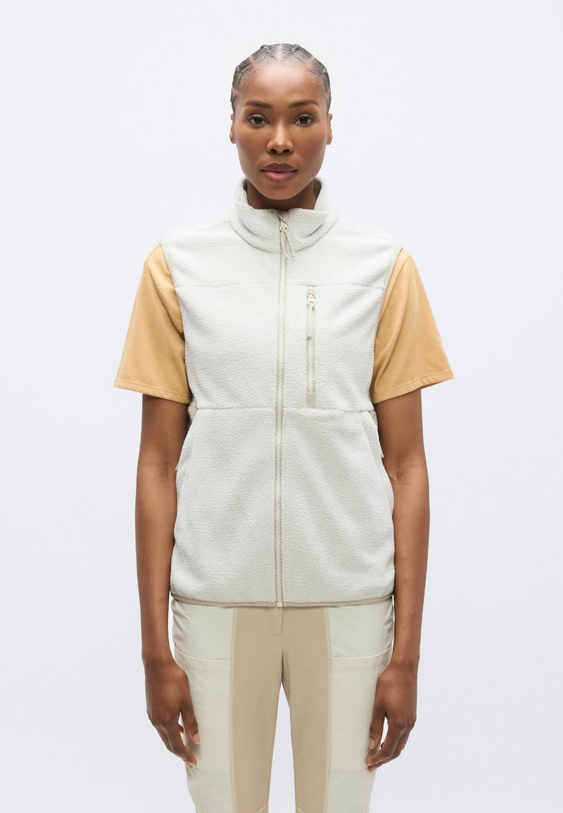 Γιλέκο από φλις σε ανοιχτό γκρι με ψηλό γιακά, μπροστινό φερμουάρ και τσέπη με φερμουάρ· συνδυασμένο με μπεζ τοπ με κοντά μανίκια και μπεζ παντελόνι.