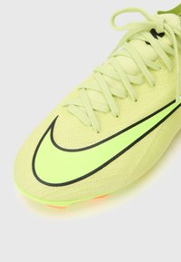 Bota de fútbol amarilla brillante con parte superior de punto texturizado, acentos negros y un logotipo de Nike prominente, con cordones planos y tacos naranjas.