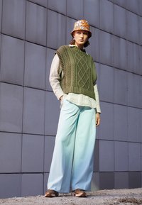 Gilet en tricot vert olive avec un motif en câble, blouse gris clair, large pantalon bleu clair et un chapeau seau à fleurs. Des baskets marron complètent le look.