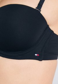 Artimas plano kadras su asmeniu, vilkinčiu juodą liemenėlę su reguliuojamomis petnešėlėmis ir maža raudona, balta bei mėlyna stačiakampio formos logo ant juostos.