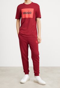 Camiseta burdeos para hombre con un logotipo rectangular en coral que dice "HUGO", combinada con pantalones jogging burdeos a juego y zapatillas blancas.