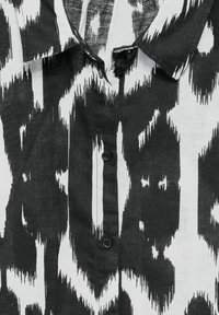 Black and white patterned shirt with a collar, featuring irregular vertical stripes and a central button closure made of fabric.