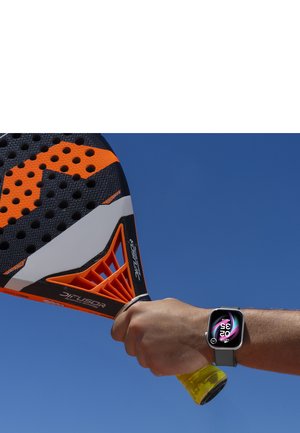 Hånd, der holder en orange og sort padelracket med blå himmel i baggrunden, iført et smartwatch, der viser tid og dato.