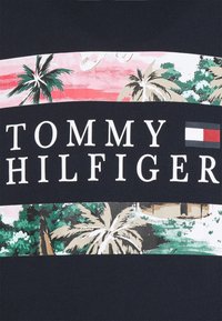 Logo Tommy Hilfiger z białymi literami i flagą w kolorach czerwonym, białym i niebieskim na ciemnej tkaninie z wzorami palm i tropikalnego krajobrazu.