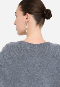 Maglione grigio a maglia con scollatura tonda, caratterizzato da accenti testurizzati sulle spalle e una finitura in tessuto liscia. Capelli lunghi raccolti in uno chignon.