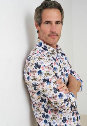 Man met gevouwen armen leunend tegen een muur, draagt een wit overhemd met een donkerblauw, roze en olijfgroen bloemmotief, kijkt naar de camera.