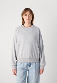 Felpa grigia con scollo rotondo, polsini e vita a coste. Presenta un logo sottile nell'angolo in alto a sinistra. Abbinata a jeans azzurri chiari.