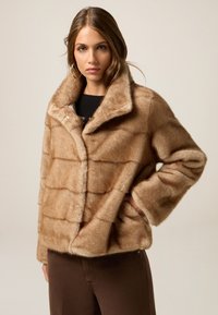 Chaqueta de pelaje sintético en marrón claro con un diseño texturizado y acolchado, que presenta un cuello alto y bolsillos frontales. Detalle de cierre a presión visible.