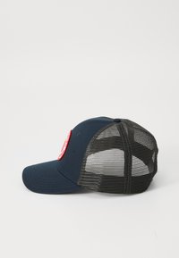 Marineblauwe en zwarte mesh trucker pet met een gebogen rand en een rode cirkelvormige patch op het voorpaneel, zijaanzicht weergegeven.